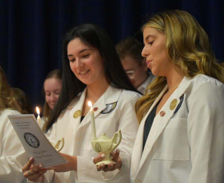 Nursing graduates in white coats holding lamps read aloud from a booklet
