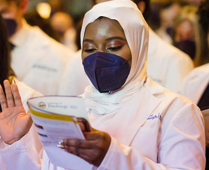 a woman in a blue mask, white coat and white head scarf holding up her right hand