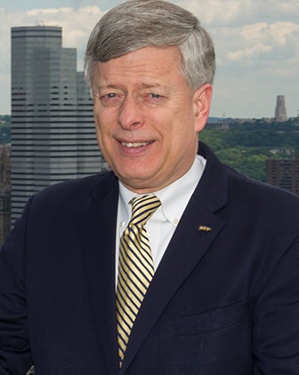 Mark A. Nordenberg in a black suit and a yellow and black striped tie stands on an overlook of Pittsburgh. 