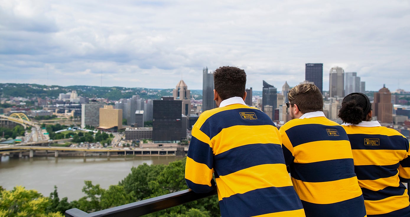 Three Pitt Pathfinder students wearing blue and gold striped rugby shirts look out over the city of Pittsburgh skyline