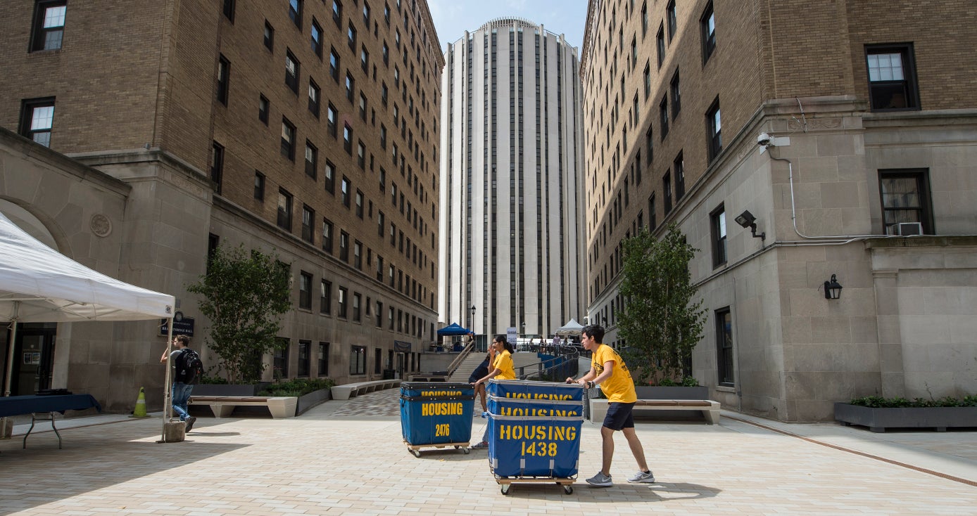 Two students in yellow shirts and blue shorts push housing cart-bins across paved plaza during move-in