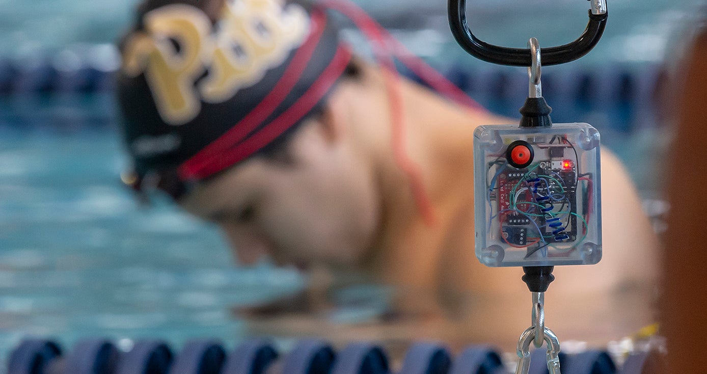 An electronic device hanging by a clip with a swimmer with a Pitt cap on in the background