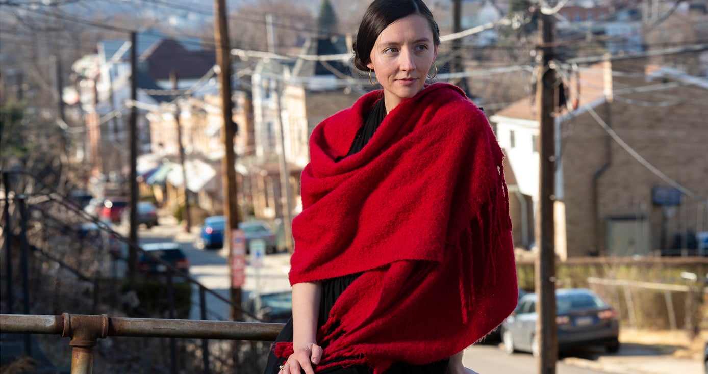 Howell draped in a red scarf, sitting on a porch ledge with a city street in the background