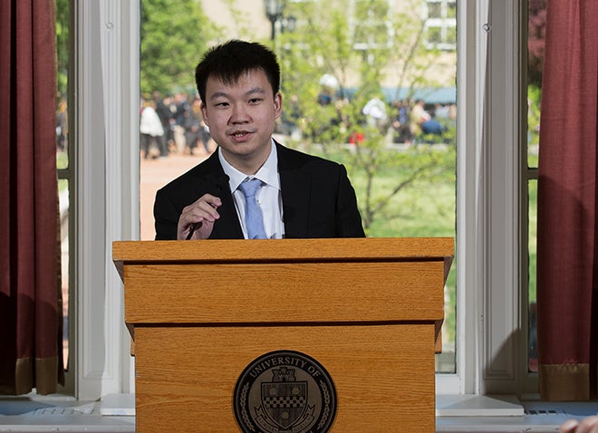 a young man in a suit at a podium with green trees and people behind him out the window