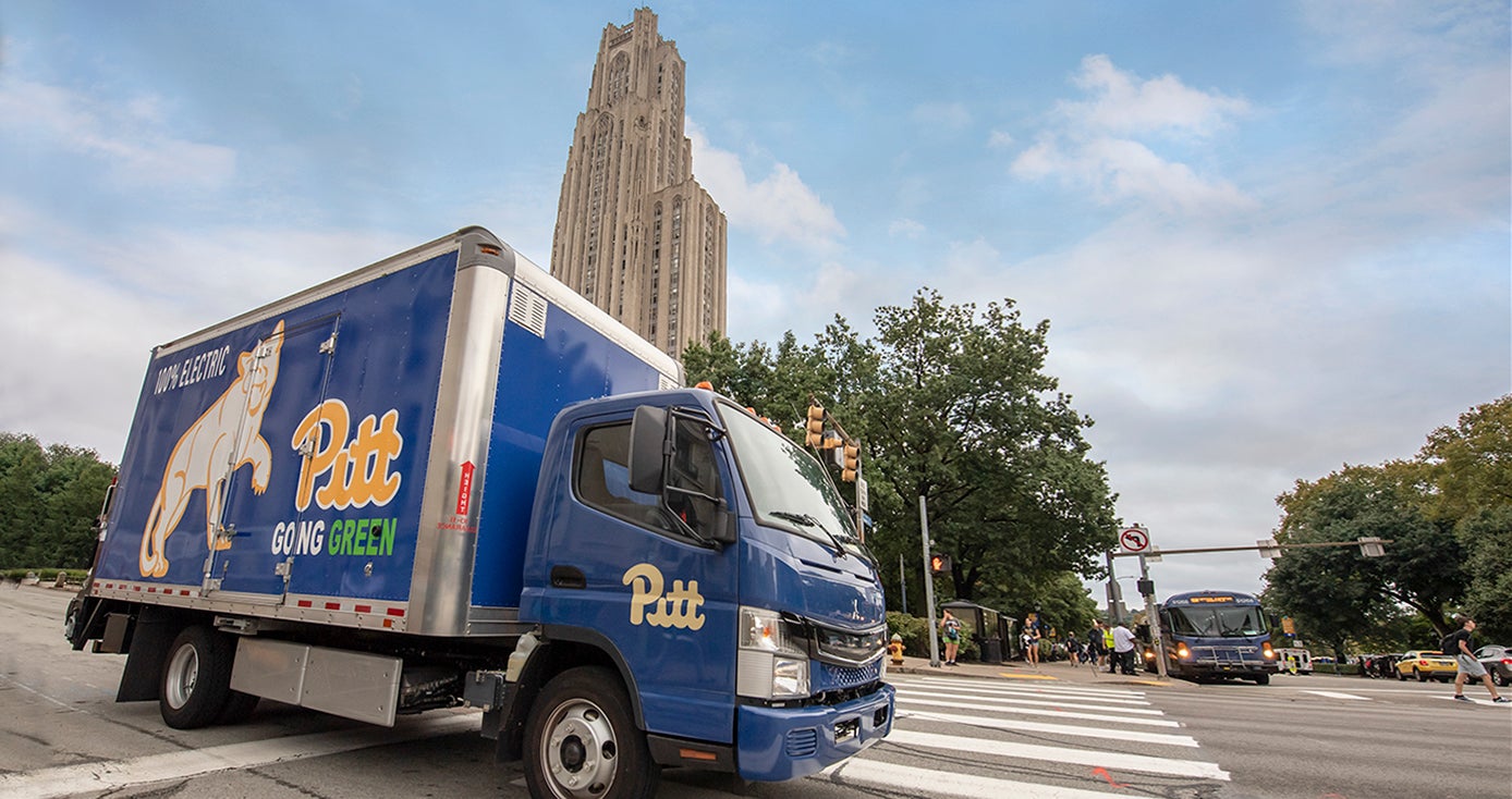 Blue box truck with panther, script Pitt logo, going green and 100% electric painted on it, with Cathedral of Learning in the background