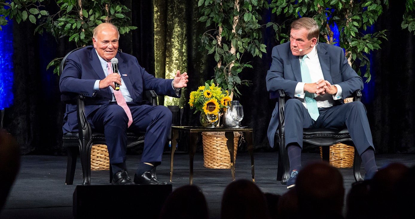 Former Pennsylvania governors Ed Rendell and Tom Ridge on stage with greenery behind them and a table with sunflowers between them. Rendell is speaking into a microphone and gesturing and Ridge's head is turned toward Rendell 