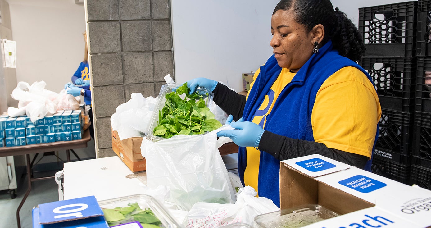A woman in a yellow shirt and blue vest packs vegetables into a plastic bag