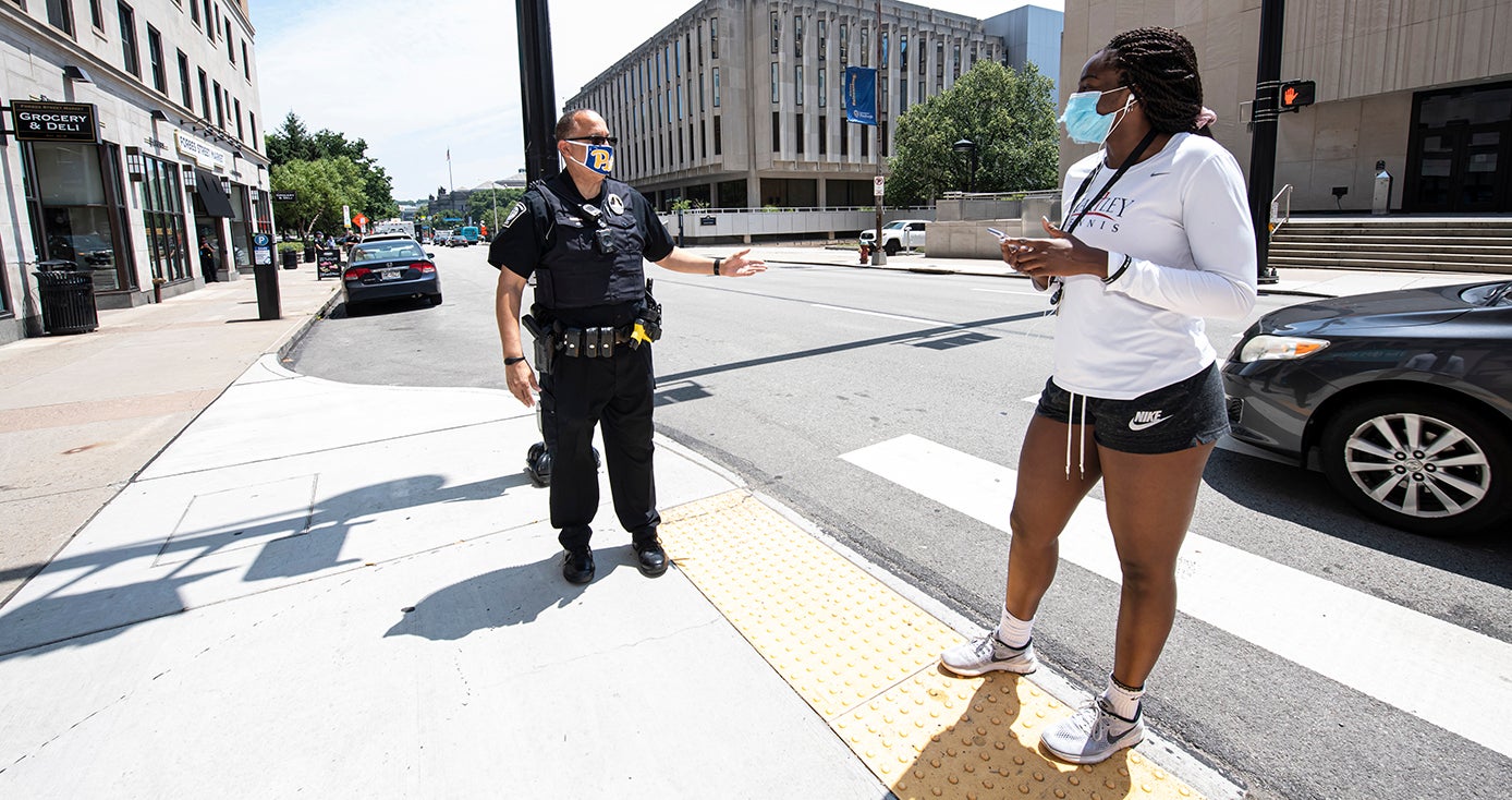 A police officer stands on a sidewalk, speaking to a person in a face mask in a white shirt and black shorts