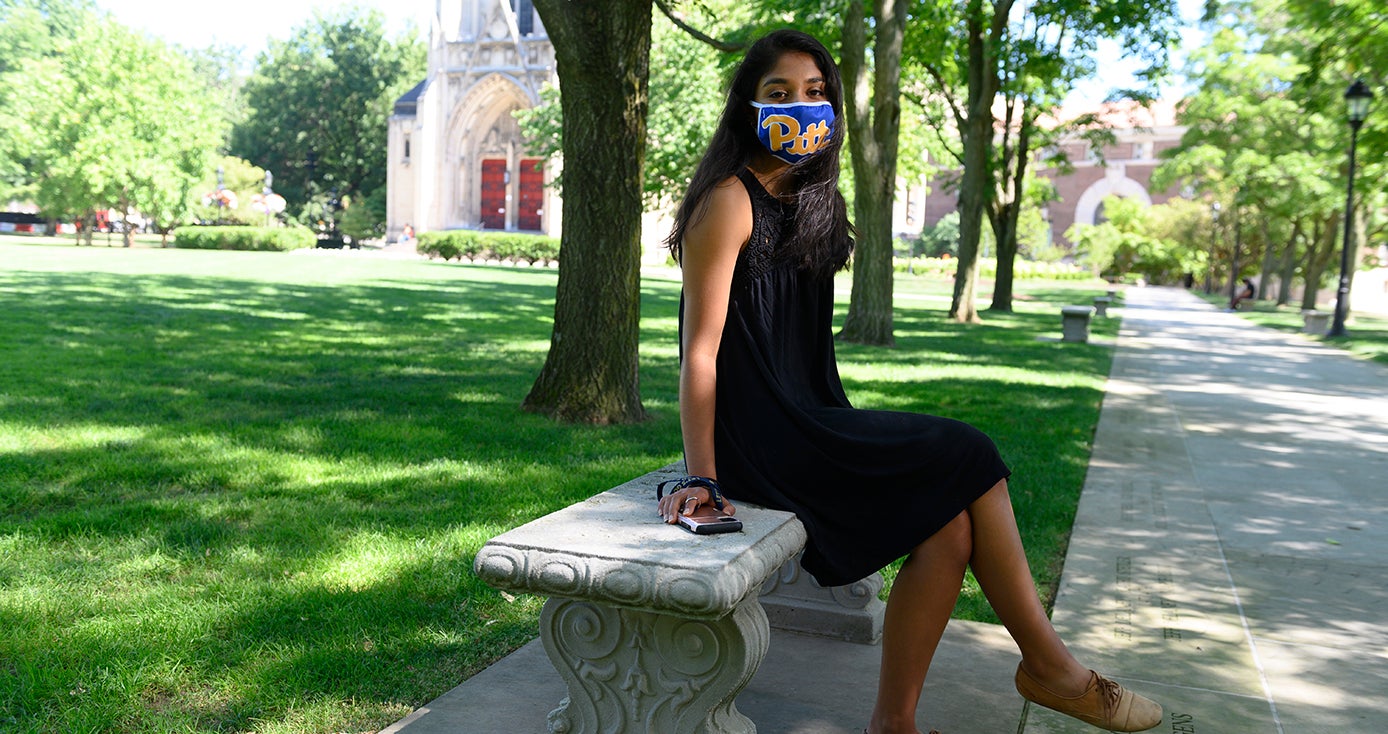 a woman in a black dress wearing a Pitt branded mask and sitting on a bench outside Heinz Chapel