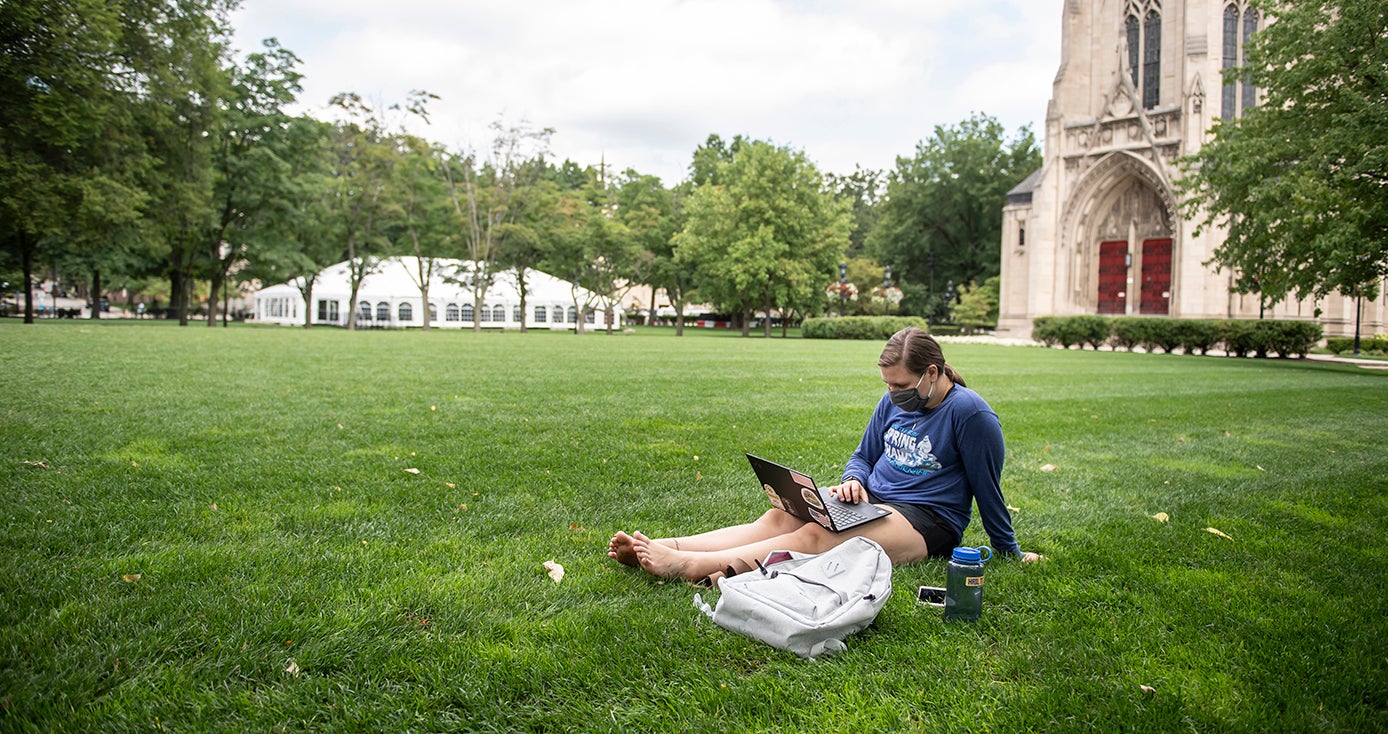 A person in a face mask uses a laptop while sitting on the lawn in front of Heinz Chapel
