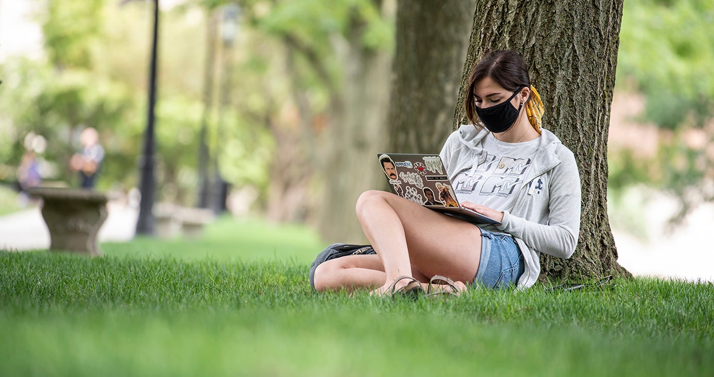 A person in a face mask, a sweater and shorts sitting by a tree, on a laptop