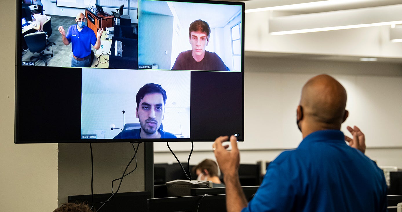 A man in a mask faces a large screen with students watching on a Zoom call