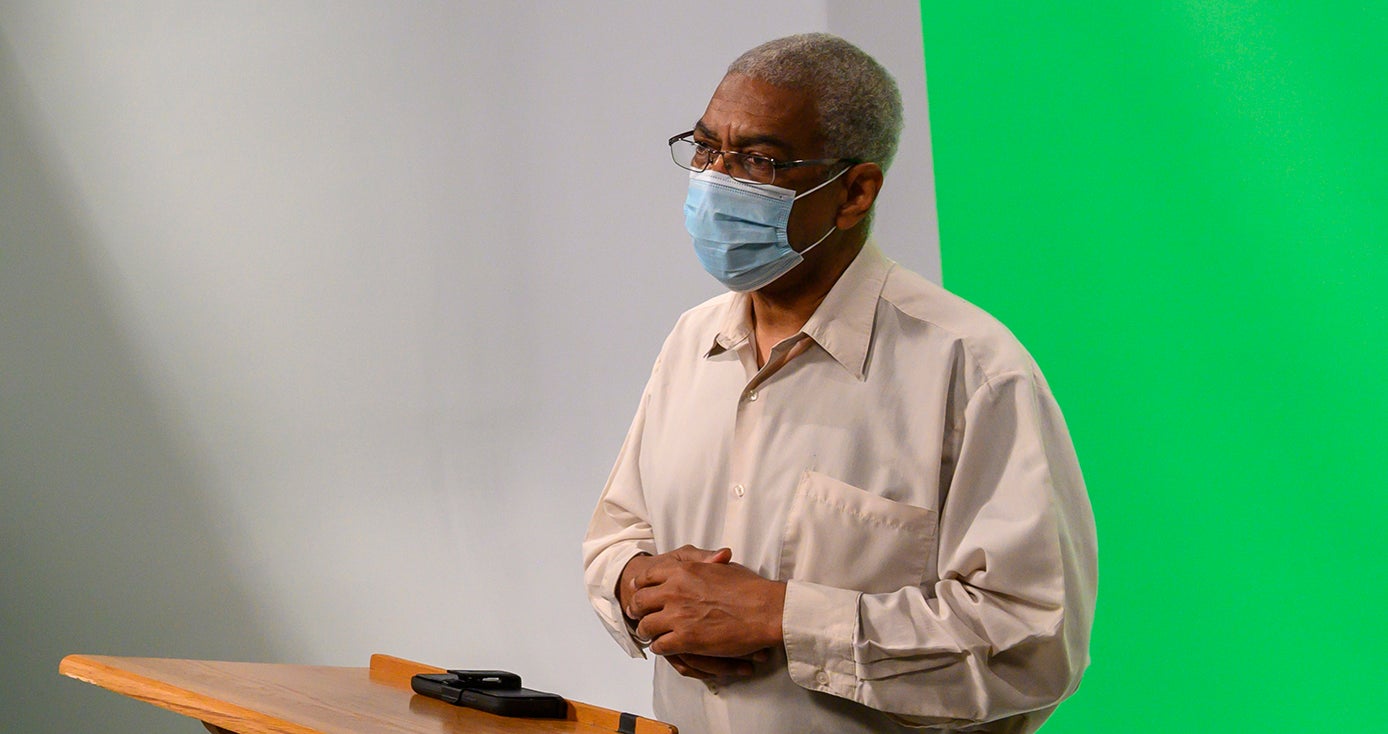 A person in a face mask and tan shirt stands in front of a green screen