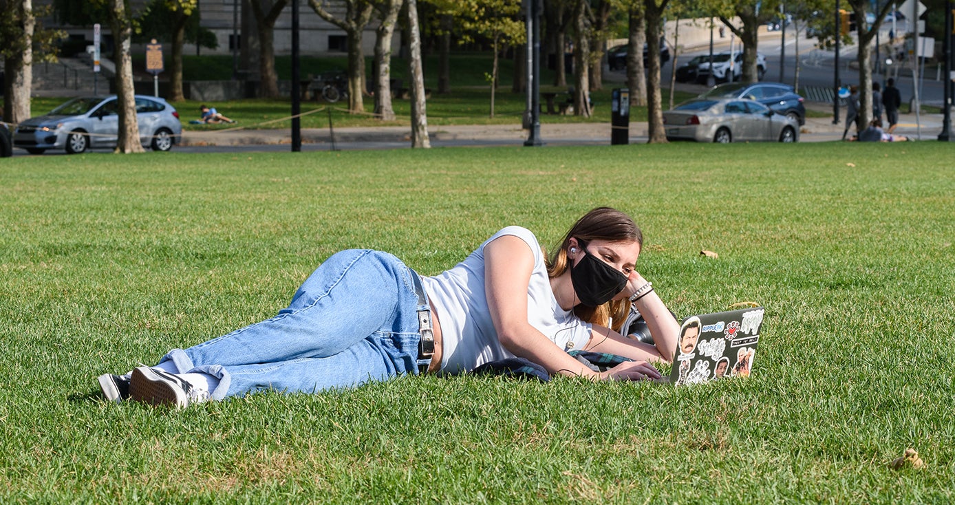 A student in a face mask on a laptop while laying down in a field