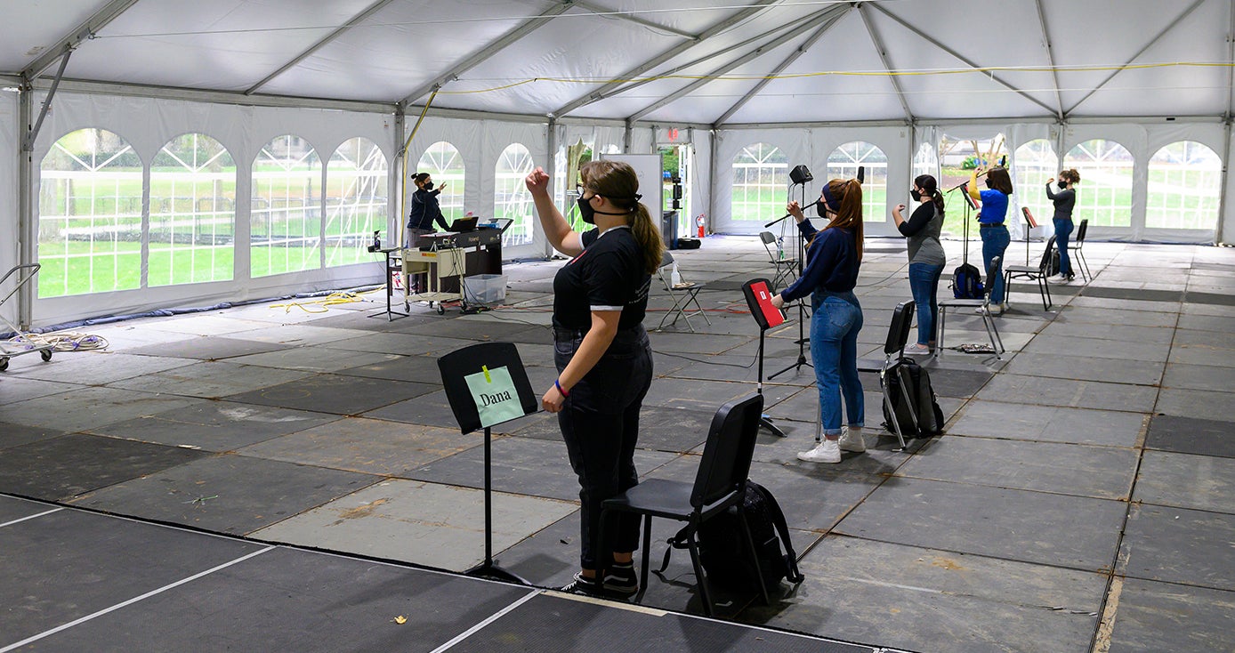 A woman in a face mask conducting to several students in face masks, inside a white tent
