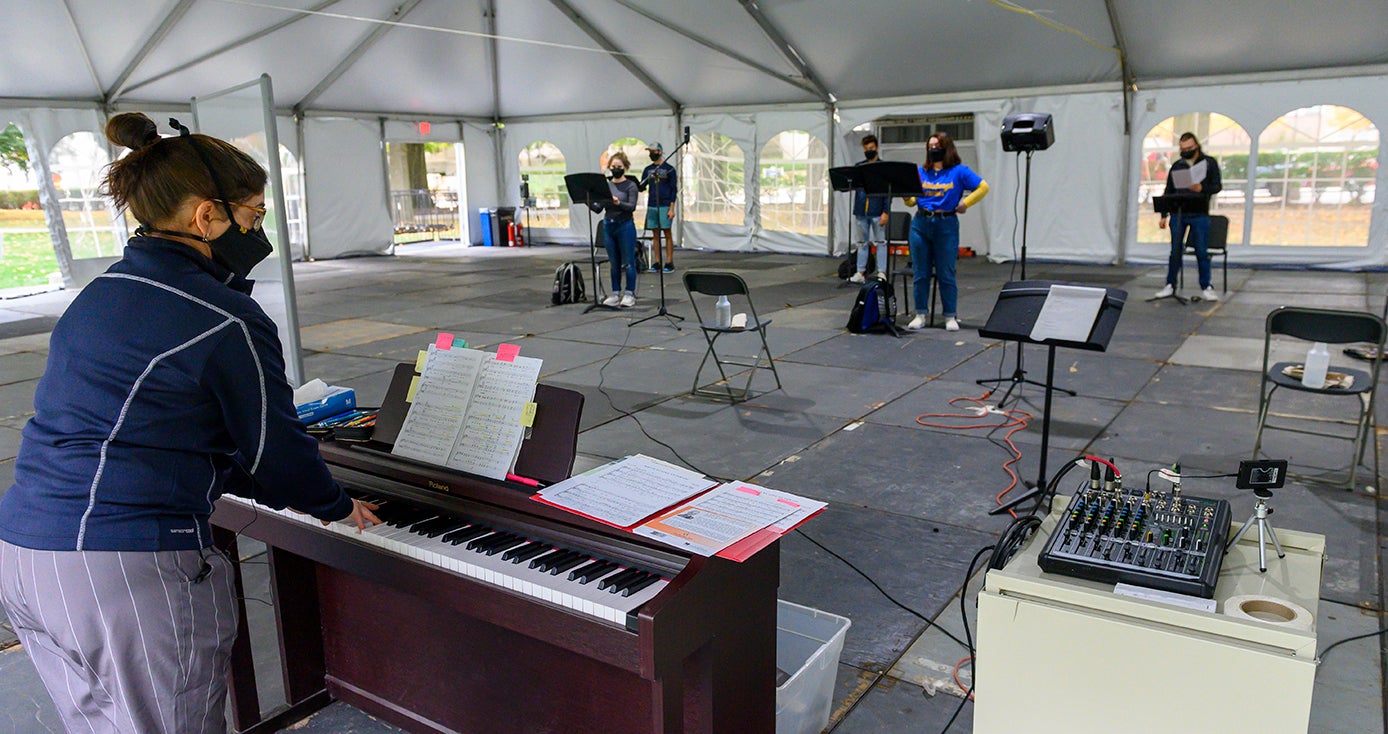 A person plays a piano facing five singers, all inside a tent 