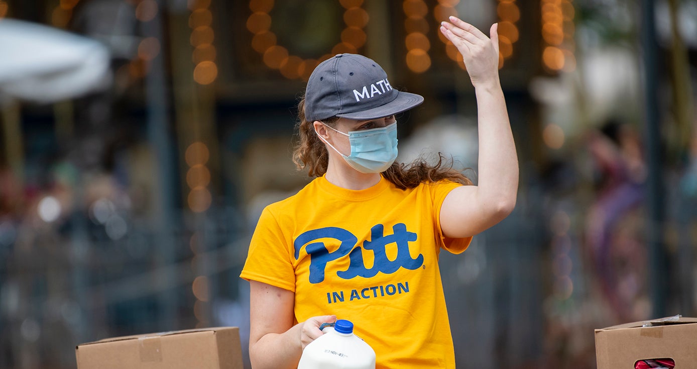 A person in a yellow Pitt shirt, gray hat and face mask waves at someone while holding a gallon of milk