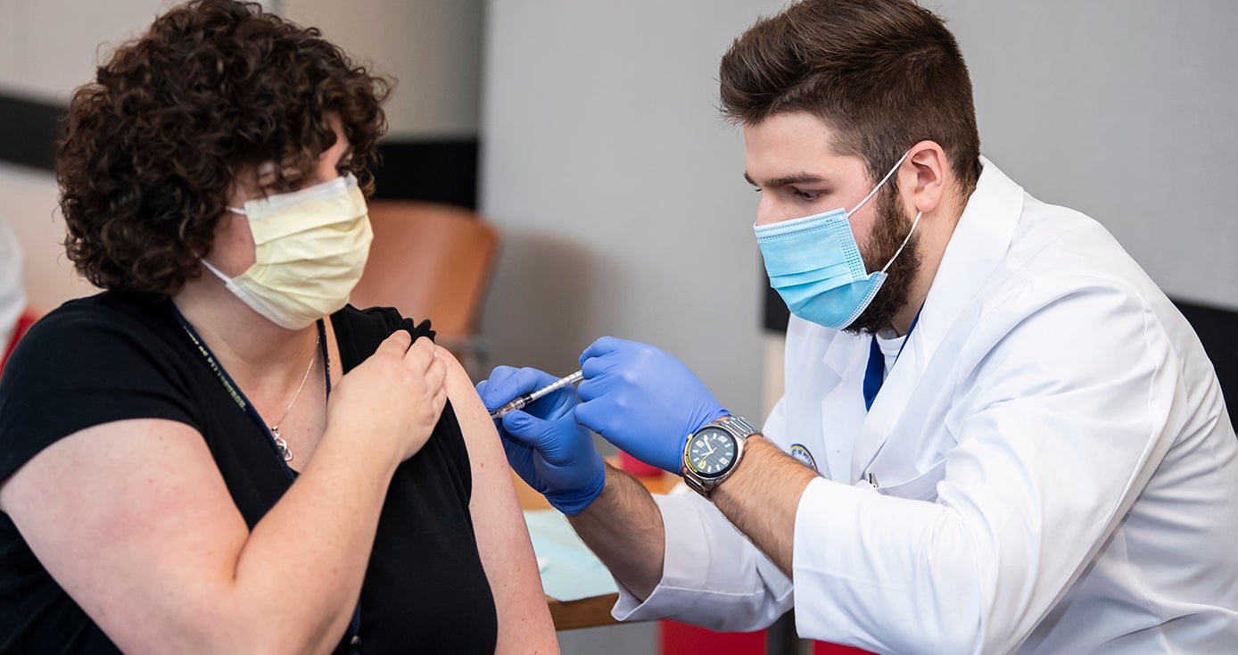 A man in a face mask and white coat and blue gloves administers a vaccine to a person in a yellow face mask and black shirt