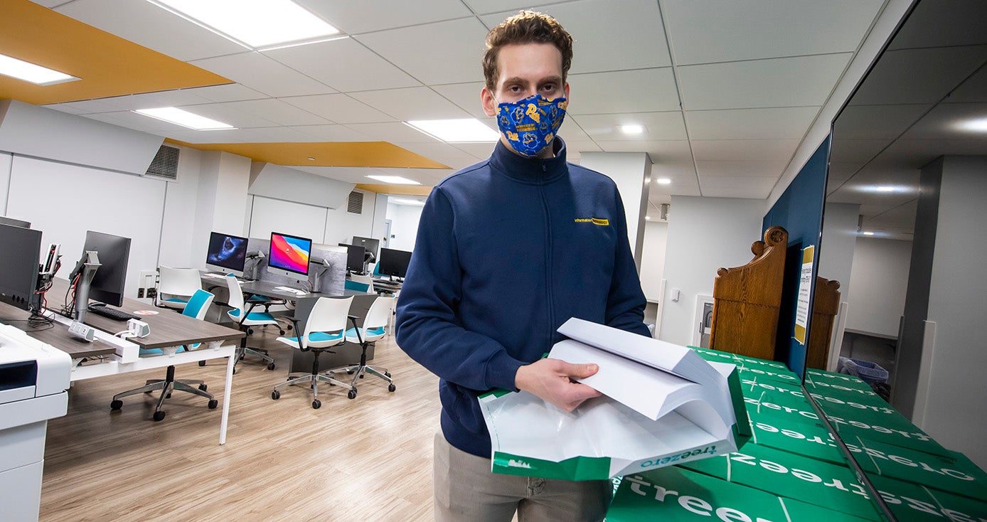 A person in a blue jacket and face mask holds a ream of paper in a classroom