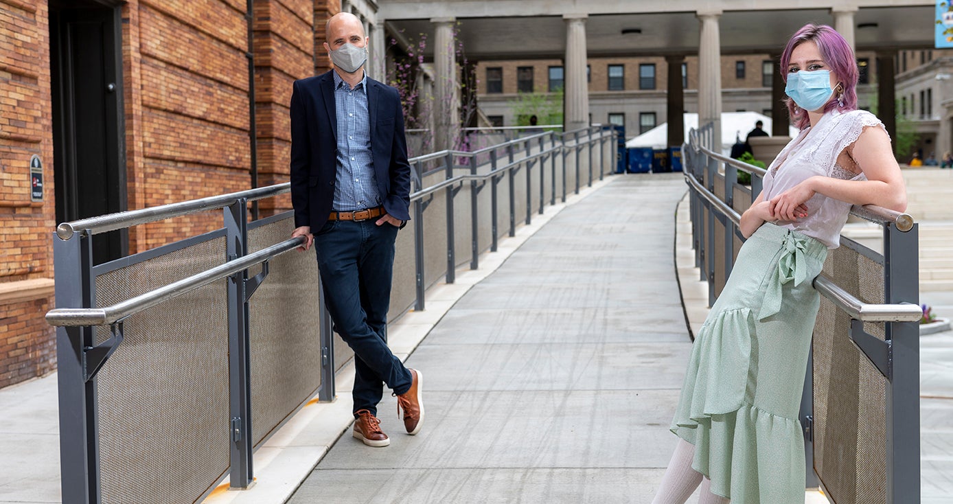 Dan Kubis leans against a rail on a walkway outside of a residence hall.  To the right is a woman wearing a mask.