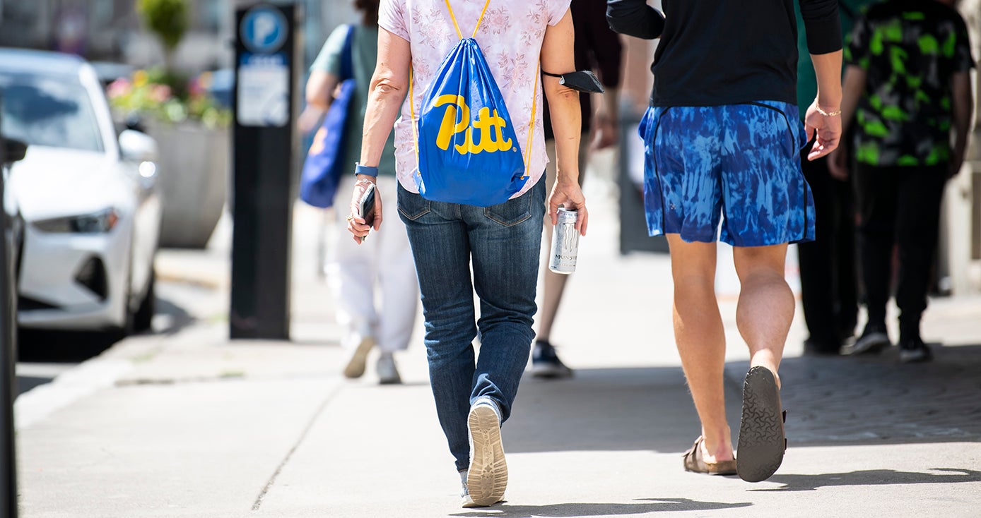 Two students walking down a sidewalk form behind, one wearing a blue Pitt bag