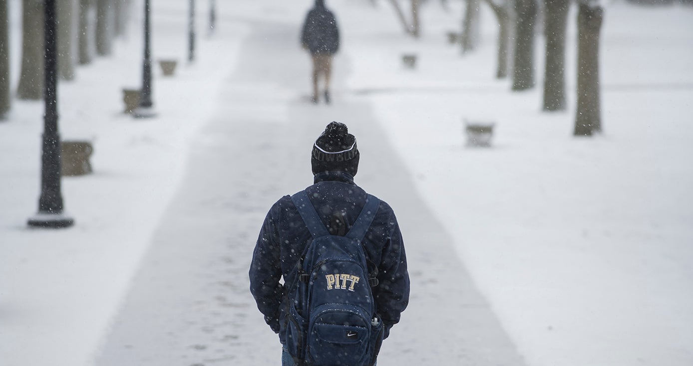 A person with a Pitt backpack and a winter hat walks away from the camera on a snowy campus
