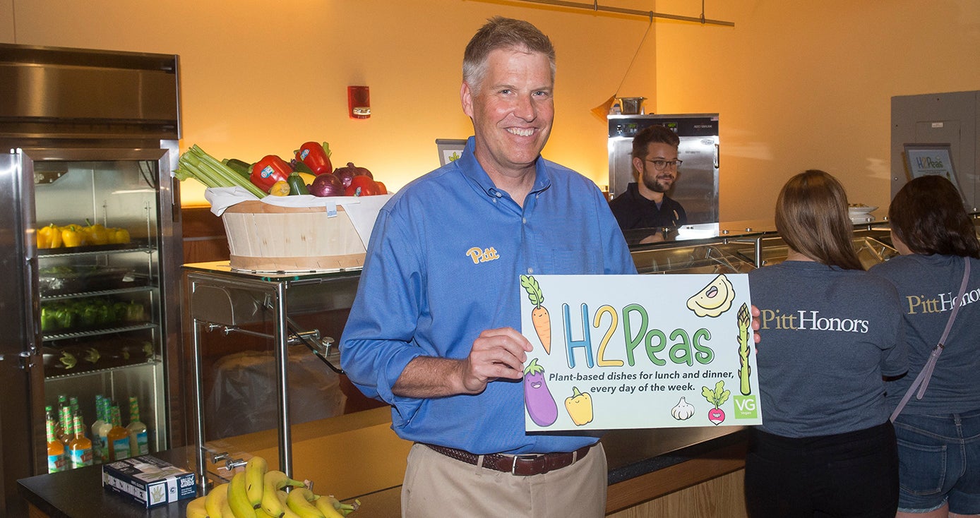 Chancellor Patrick Gallagher holding an H2Peas sign decorated with images of vegetables, standing in front of a cafeteria station with fresh food on display and customers in line for food