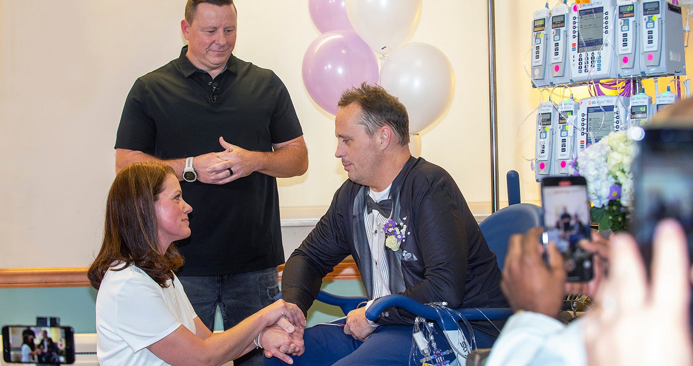 Matt Shilling sitting in a chair in a hospital room, connected to various medical equipment, while Amy Harvey kneels, holding his hands, and their pastor looks over them during a wedding ceremony
