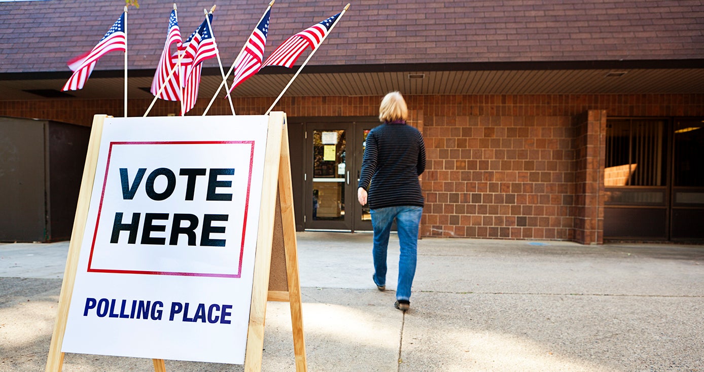 Brick building in background with person walking toward it a "Vote Here' sign in the foreground with American flags on top of it