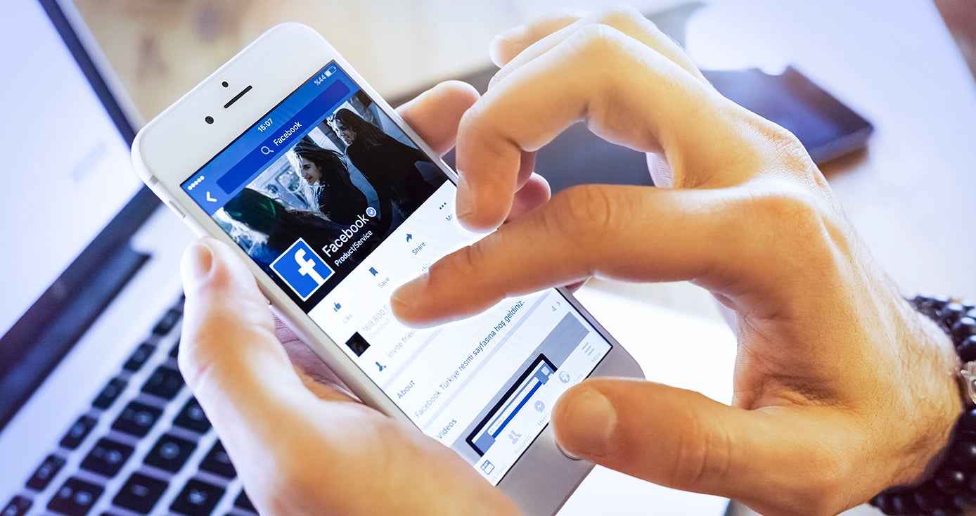 Close up of a person's hand holding a smart phone displaying Facebook, with a laptop computer in the background. 