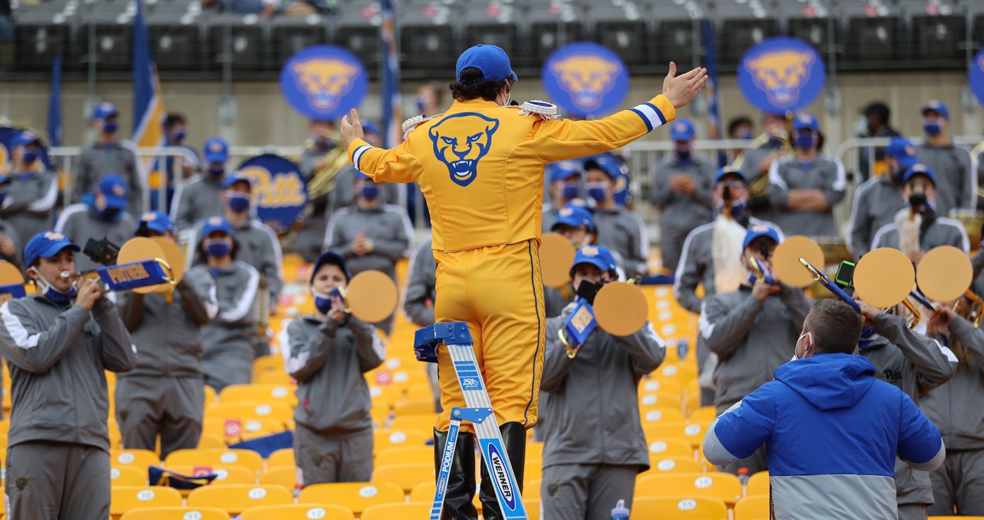 A person in yellow Pitt gear raises their hands in front of a band