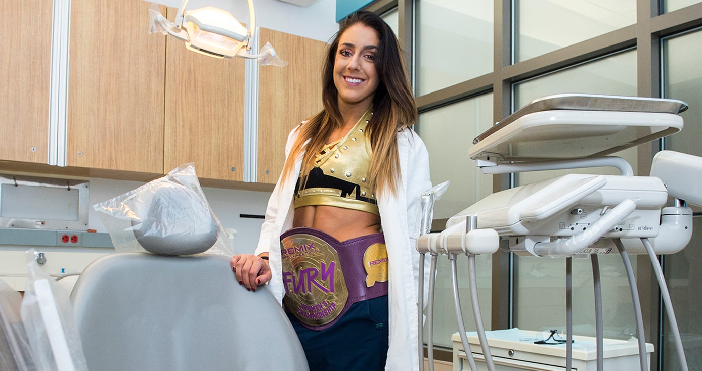 Britt Baker, a woman with long brown hair, wearing a white dentist's coat and a wrestling belt