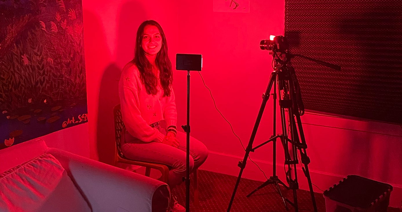 A person sitting in the corner of a dark red room with a camera facing them