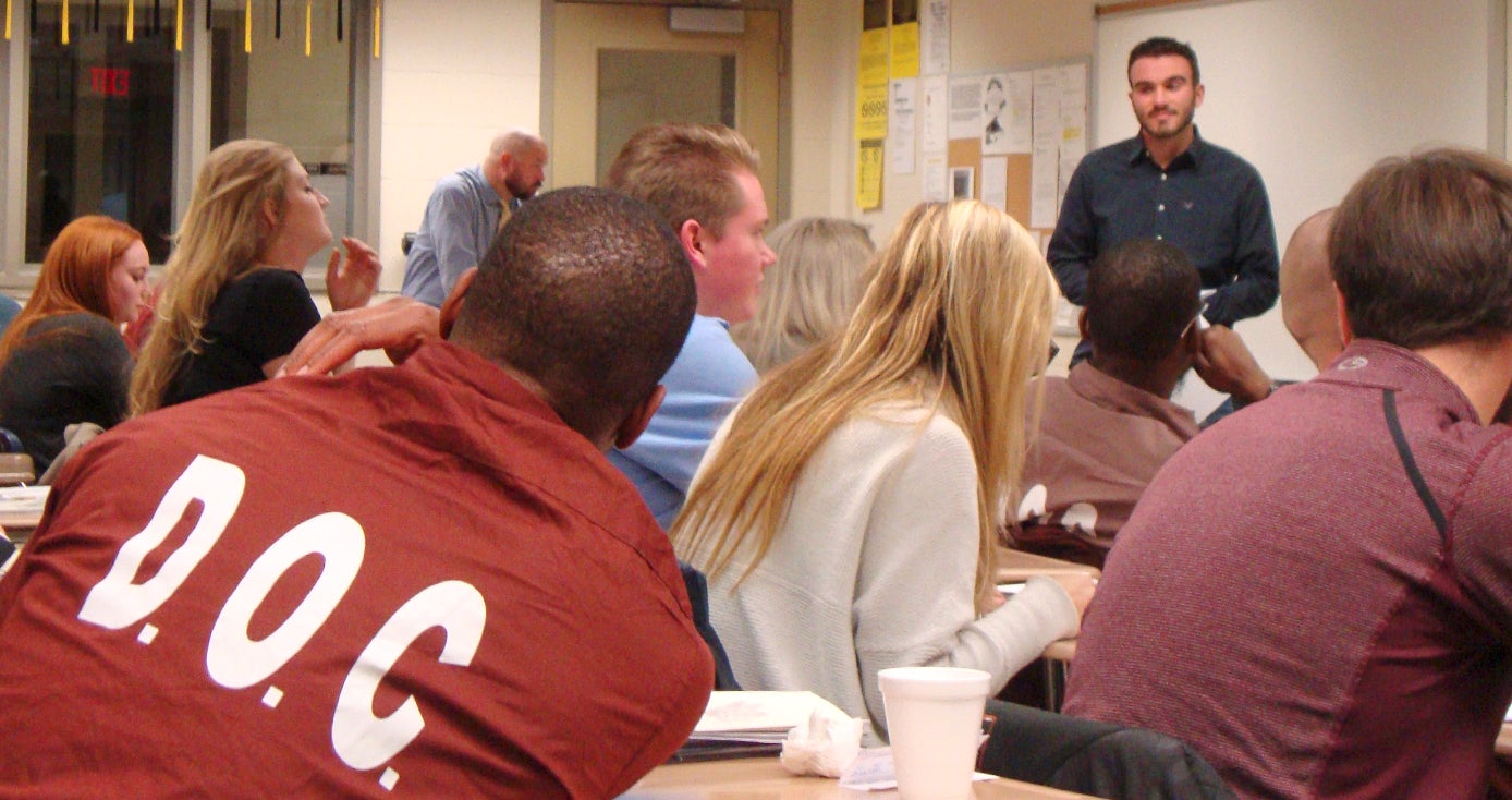 Multiple students with backs facing the camera with professor in background leading class. Student closest to camera has DOC on back of prison uniform.