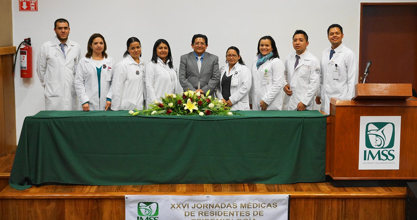 Nine people in white coats stand for a picture in front of a green table with a floral arrangement