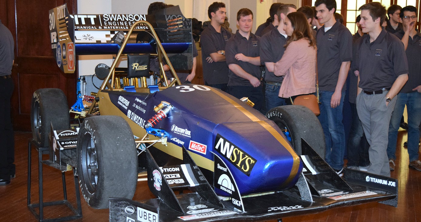 racecar in a blue and gold color theme with a group of students and onlookers standing on the right side of photo