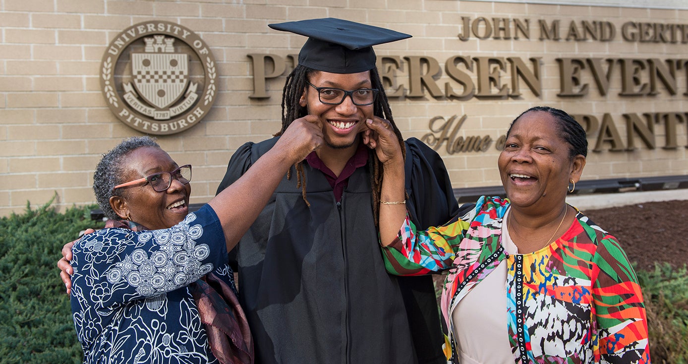 male graduate in cap and gown; his mother and aunt on either side of him pinching his cheeks