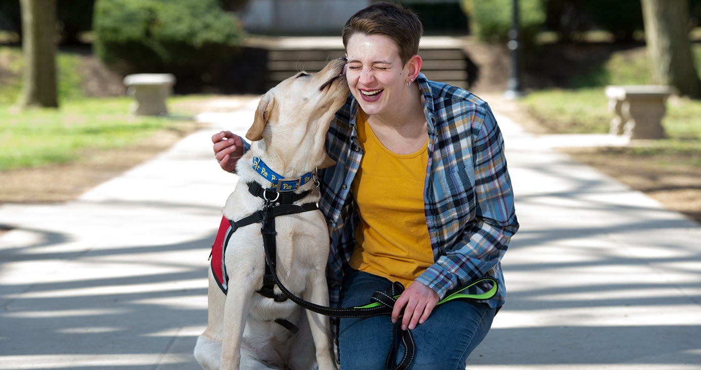 a light tan lab-looking dog licking the check of a student in a yellow shirt and blue plaid overshirt