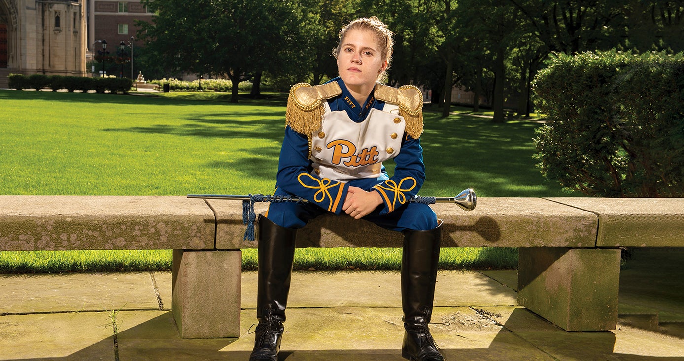 Crissy Shannon in blue and gold PItt marching band drum major uniform sitting on cement bench with baton across her lap.