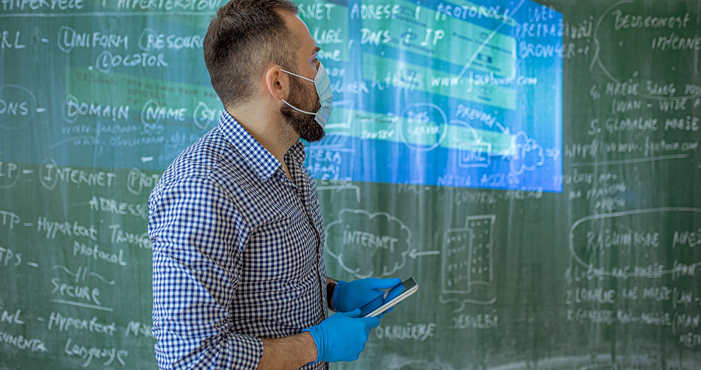 a teacher in a mask and gloves holding a tablet in front of a chalkboard