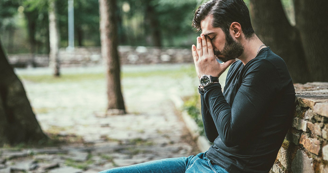 a man in a longsleeve shirt and jeans sitting outside with his hands on his face and his eyes closed