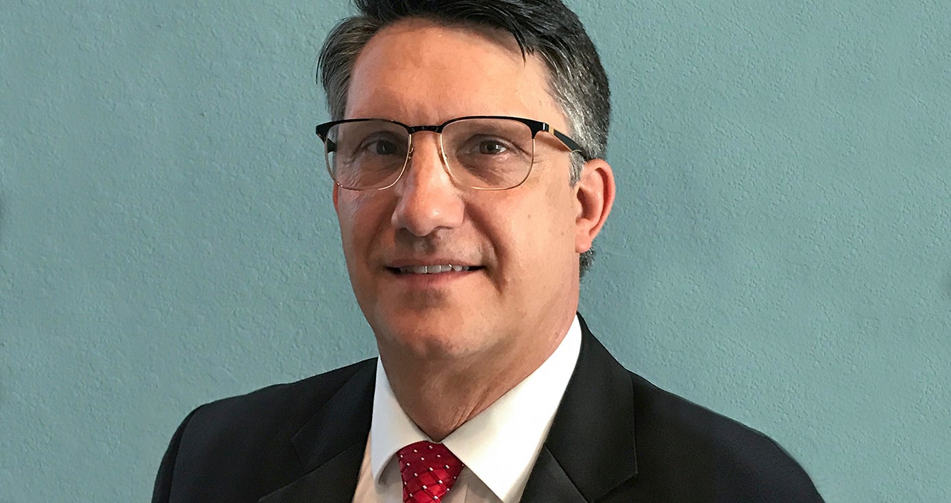 Gregerson in a dark suit, white shirt and red tie in front of a teal background