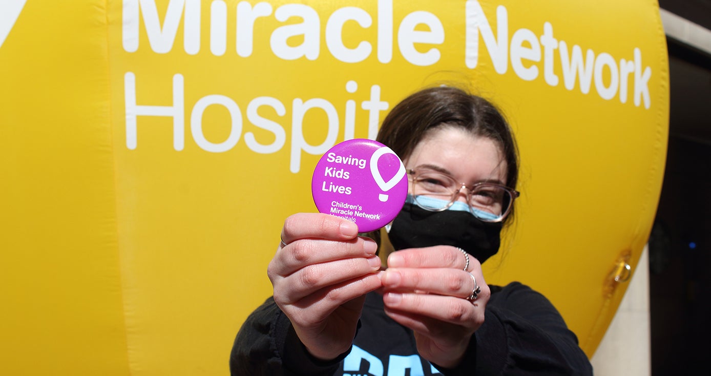 a person in a black sweatshirt and black mask holding a pink button that says "Saving Kids Lives"