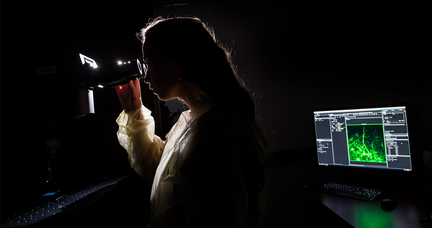 A backlit woman looks into a scope with a computer monitor behind her displays a glowing green image
