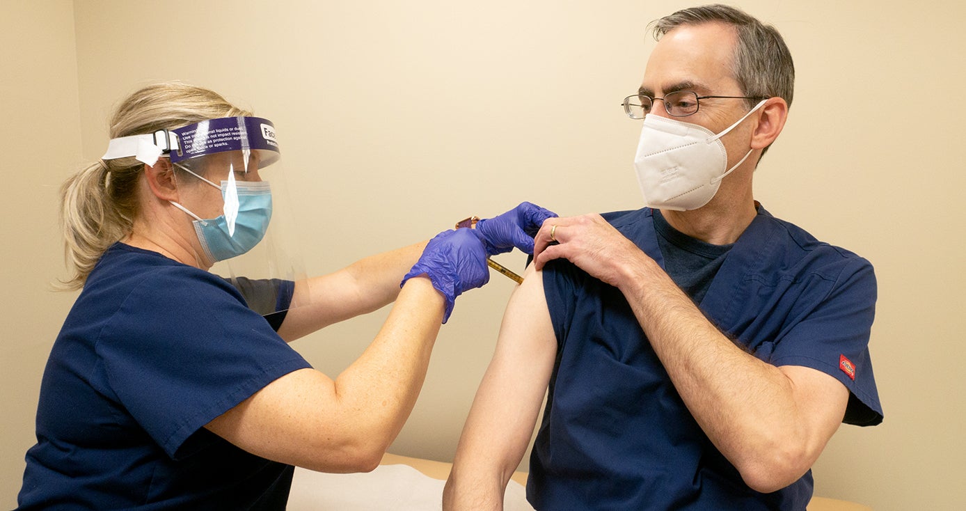 A physician with a face mask and shield gives another person in a face mask a shot