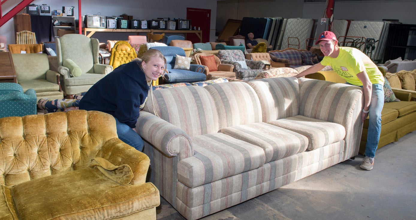 two students, a woman and a man, wearing neon yellow shirts and moving a couch