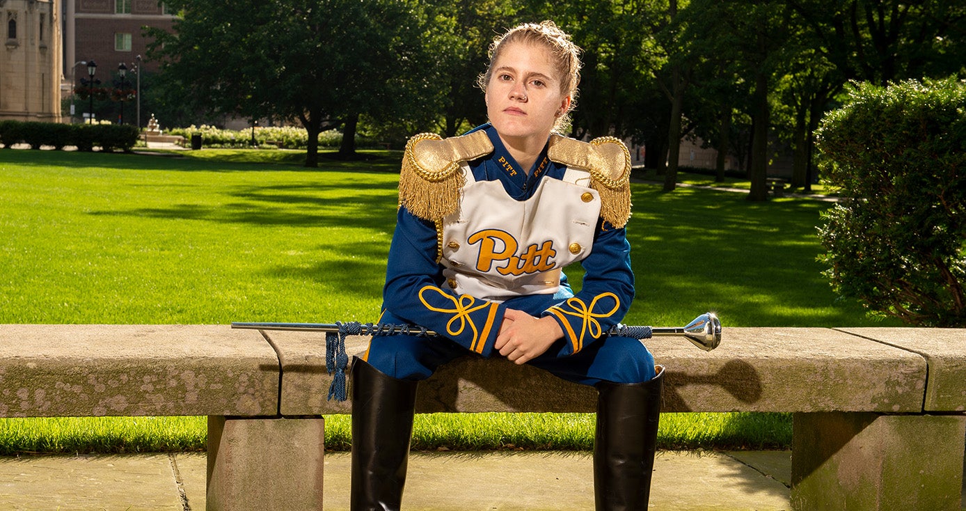 a woman in a Pitt band uniform sitting pensively on a bench outside the Cathedral of Learning
