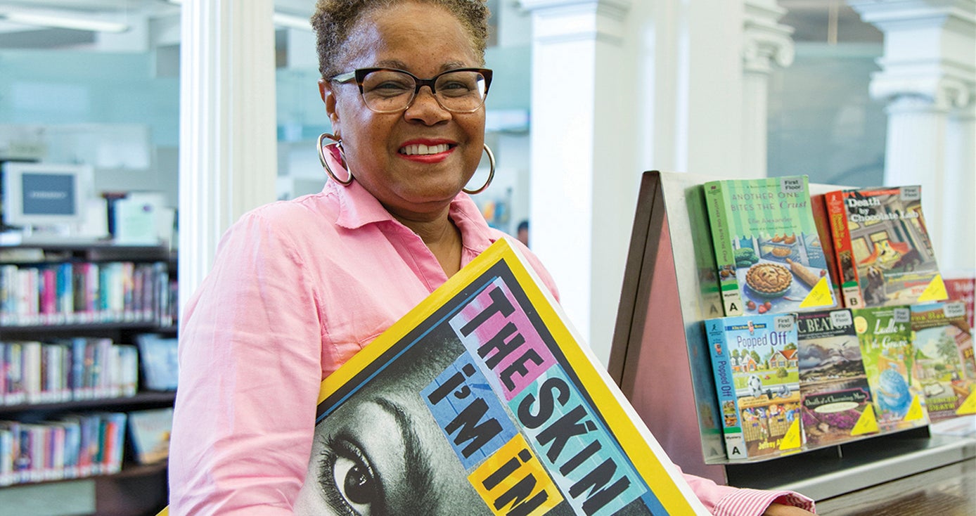 Sharon Flake, holding oversized copy of her book "The Skin I'm In," standing in a library with shelves of books in the background