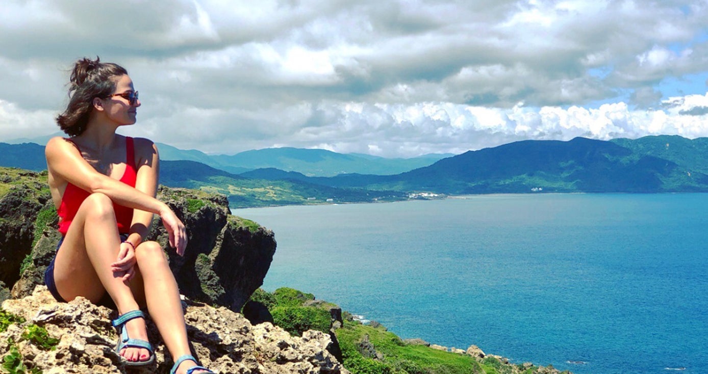 Juliette Rihl pictured seated in shorts and tank top in front of a view of mountains, water and cloudy sky