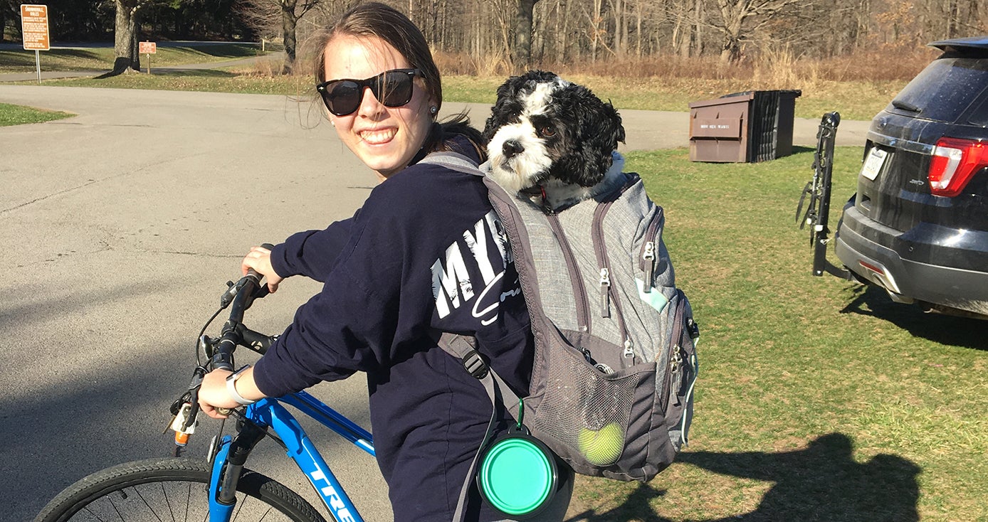 Katie Kniess with sunglasses on bicycle with Oliver, a black and white dog, in her backpack